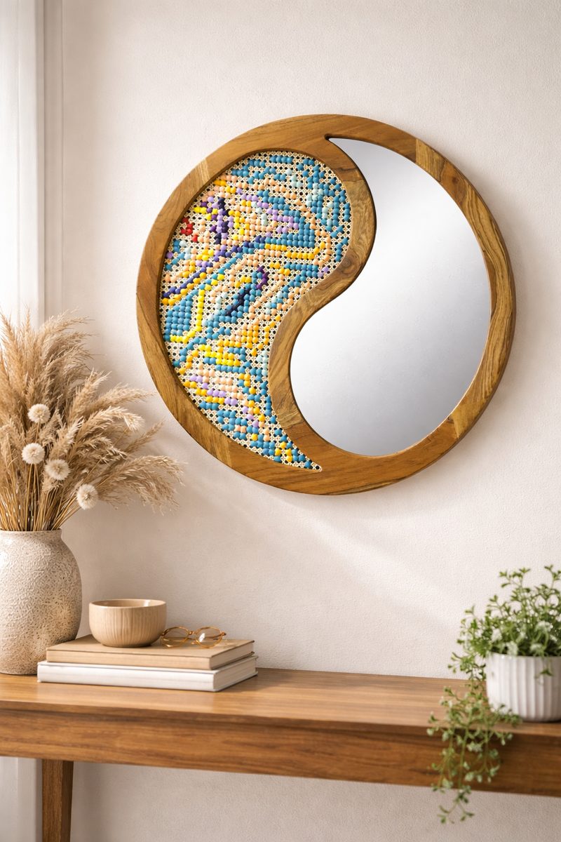 A round wooden wall mirror split in a yin and yang curve, one half a mirror and the other half thread on cane in yellow, blue and green.