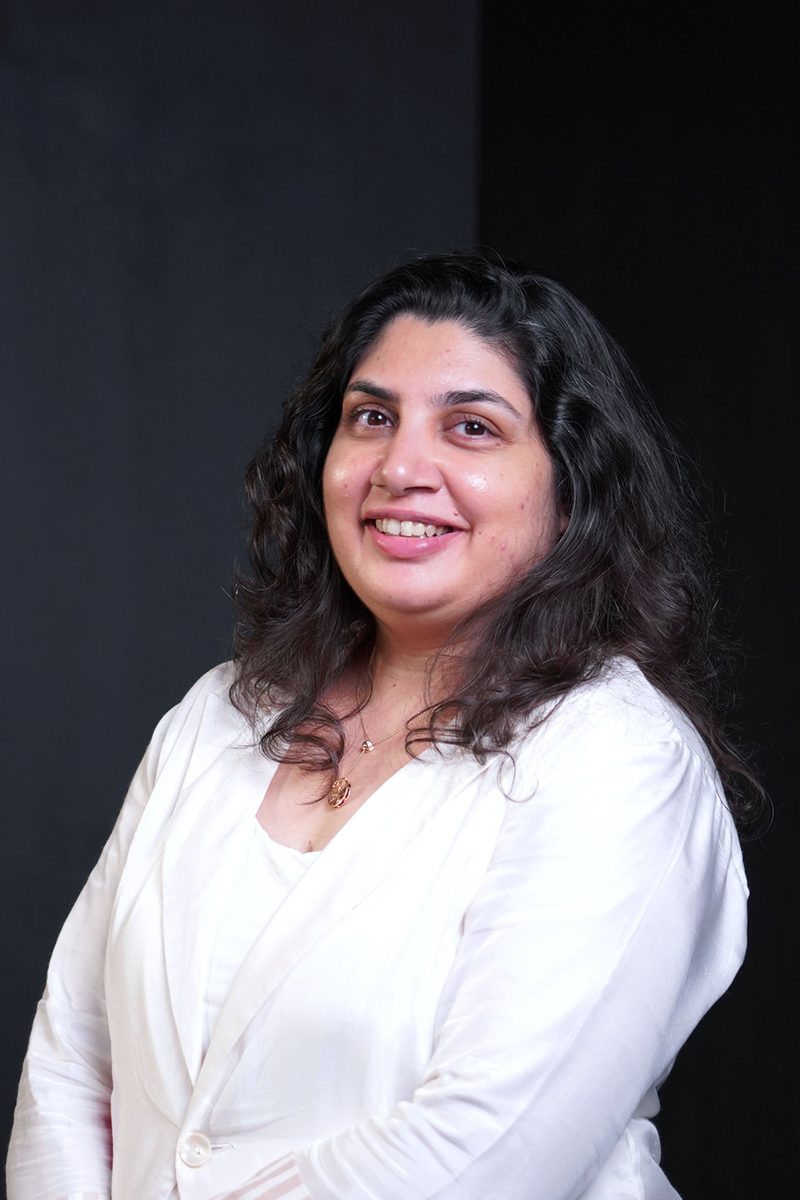Portrait of Saumya Kashyap in a white jacket, smiling, against a dark backdrop.